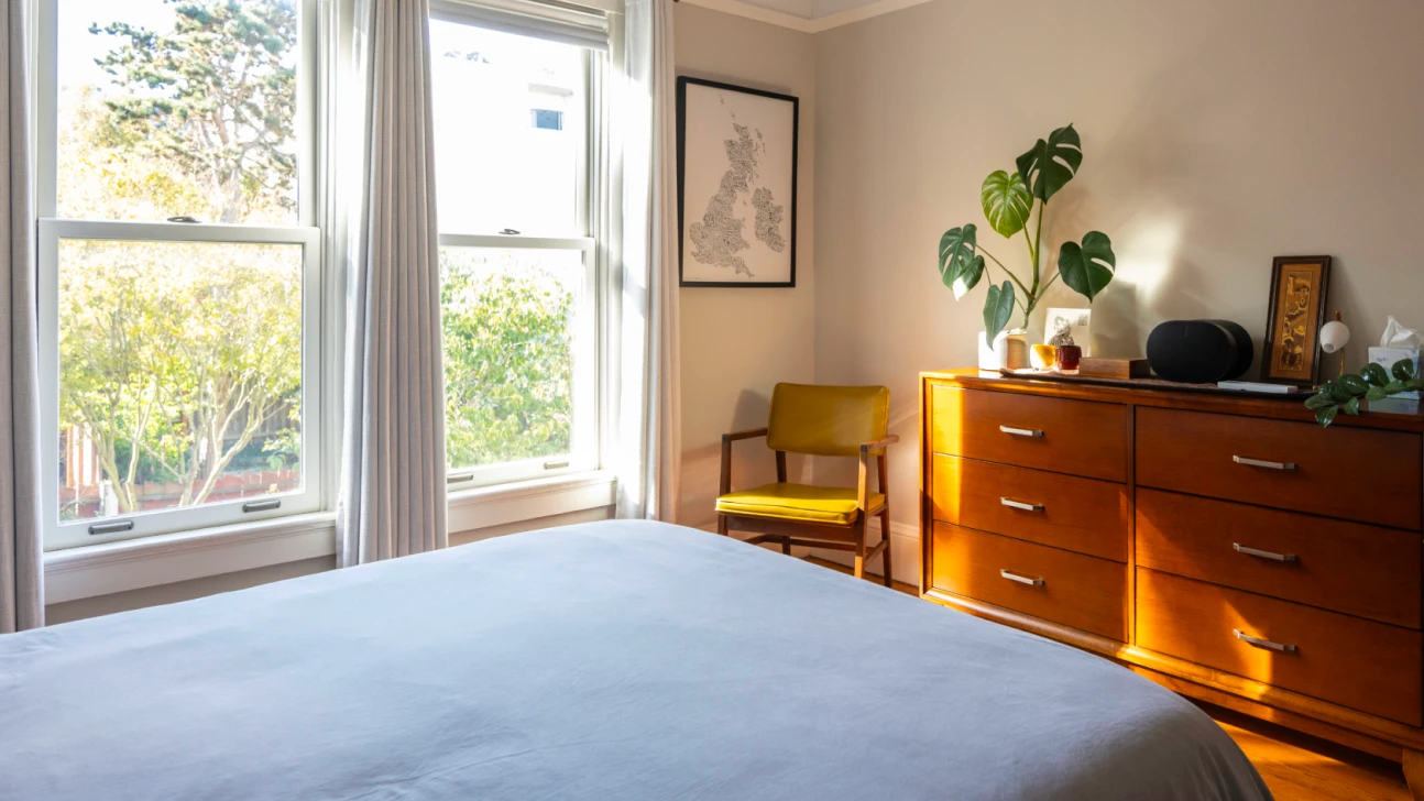 Bright Bay Area bedroom renovation with wood dresser, indoor plants, soft neutral palette, and large sunlit windows