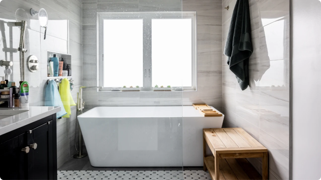 Bay Area bathroom renovation featuring freestanding soaking tub, glass shower, gray tile walls, and wood bench with natural light window