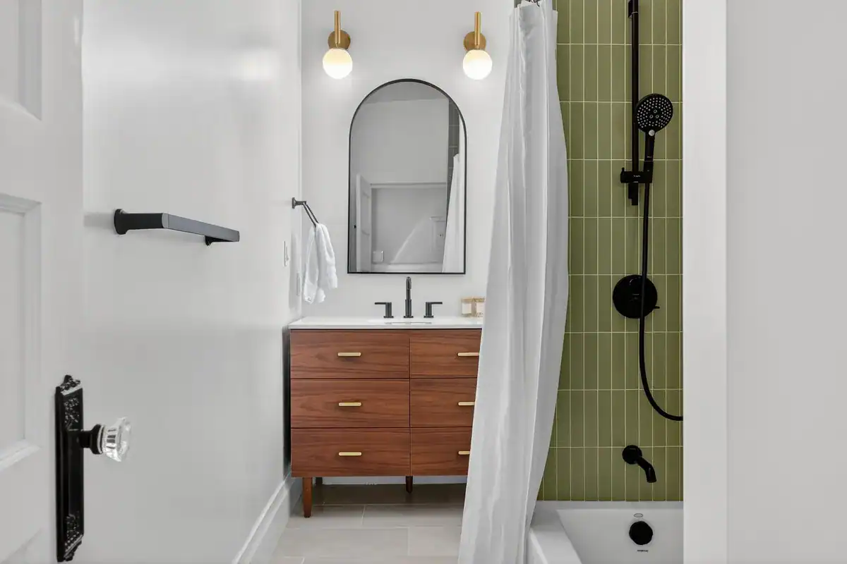 Midrange bathroom remodel with green vertical tile tub-shower, walnut vanity, arched black mirror, and brass sconces.