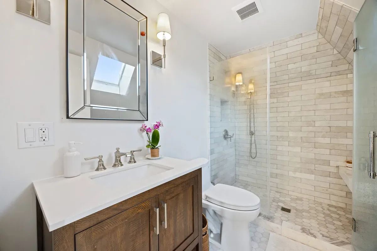 Small attic bathroom under a sloped ceiling with skylight, marble-tiled walk-in shower, hexagon floor tile, and wood vanity.