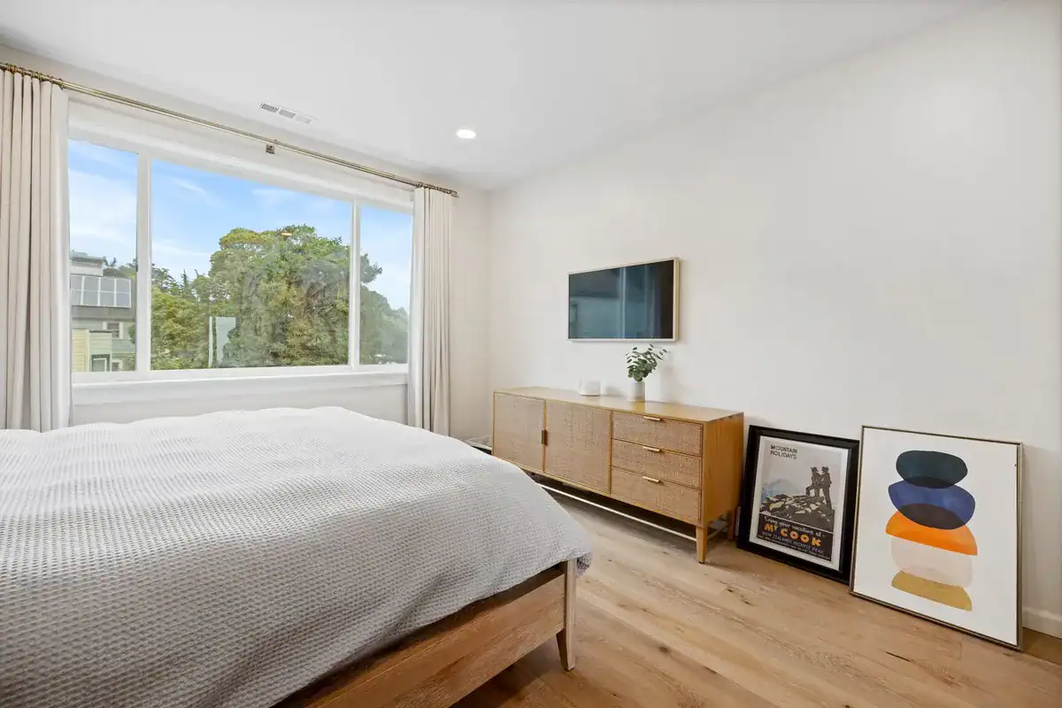 Bedroom with a wide picture window, white oak floors, recessed lighting, and a rattan-front credenza below a wall-mounted TV.