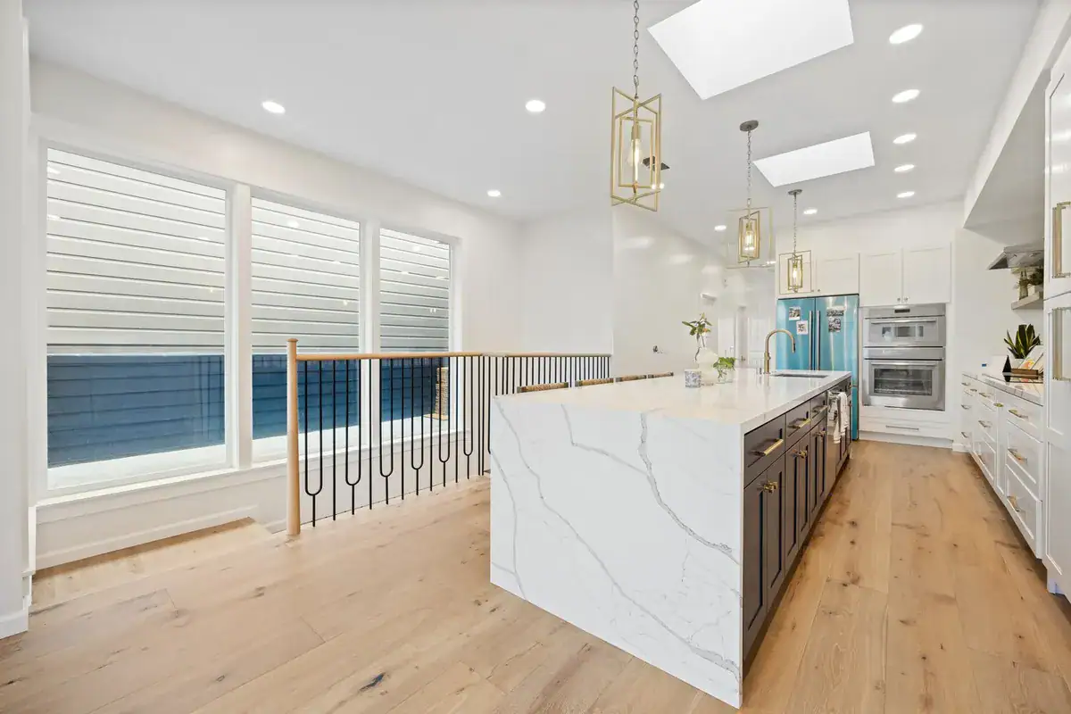 Kitchen with a white quartz waterfall island, dark lower cabinets, brass pendant lights, skylights, and wide-plank white oak floors beside an open stairwell.