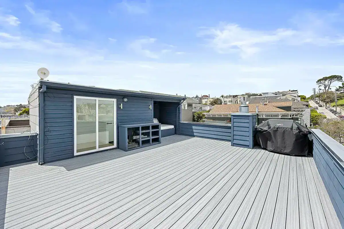 Rooftop deck with gray composite decking, blue horizontal siding, and a roof access structure with a sliding glass door, overlooking San Francisco homes.