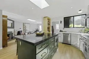 Remodeled open-concept kitchen with a green island, dark stone countertop, and skylights, opening into the living room.