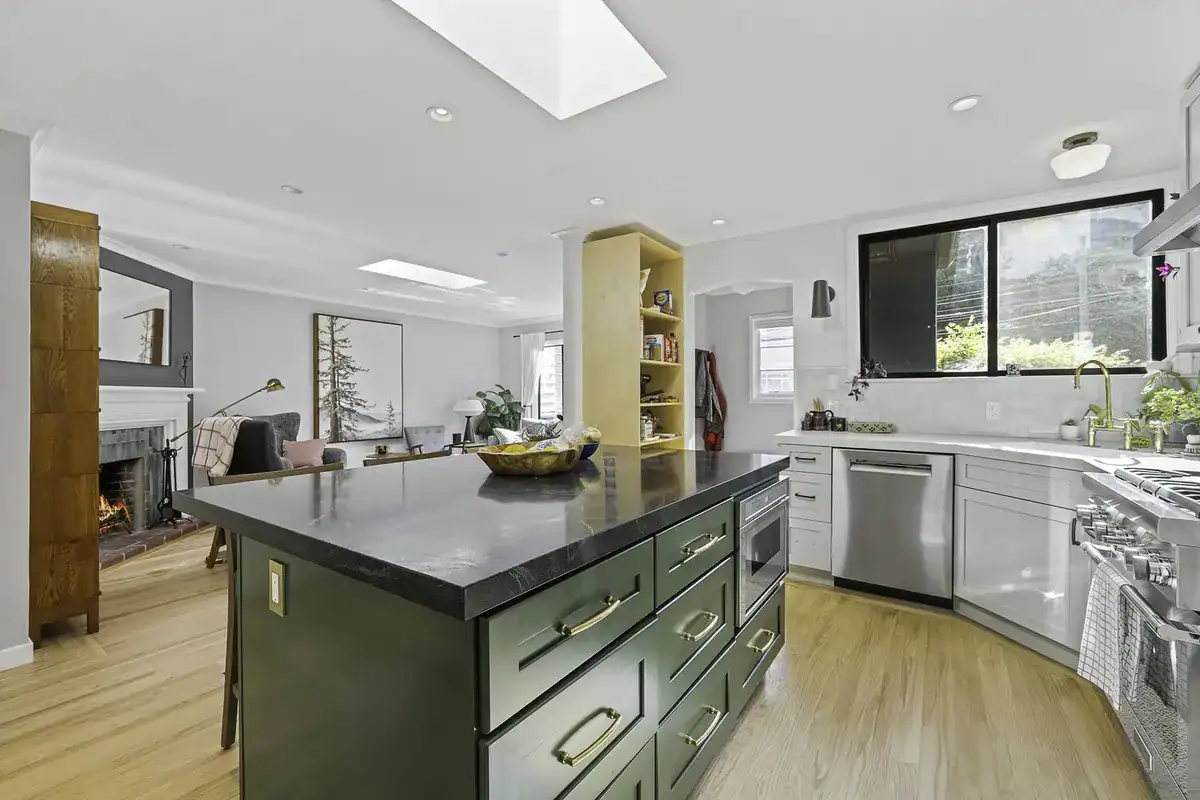Remodeled open-concept kitchen with a green island, dark stone countertop, and skylights, opening into the living room.