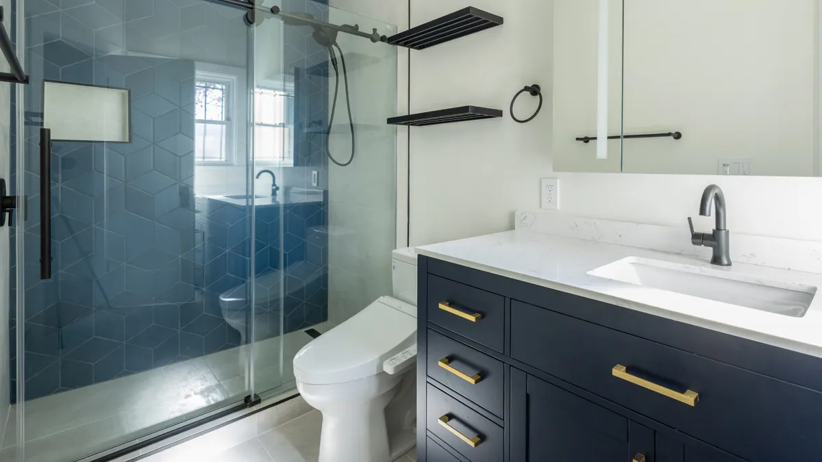 Bathroom with a blue hexagon-tile walk-in shower behind a sliding glass door, a smart toilet with bidet seat, and a navy vanity with brass pulls.