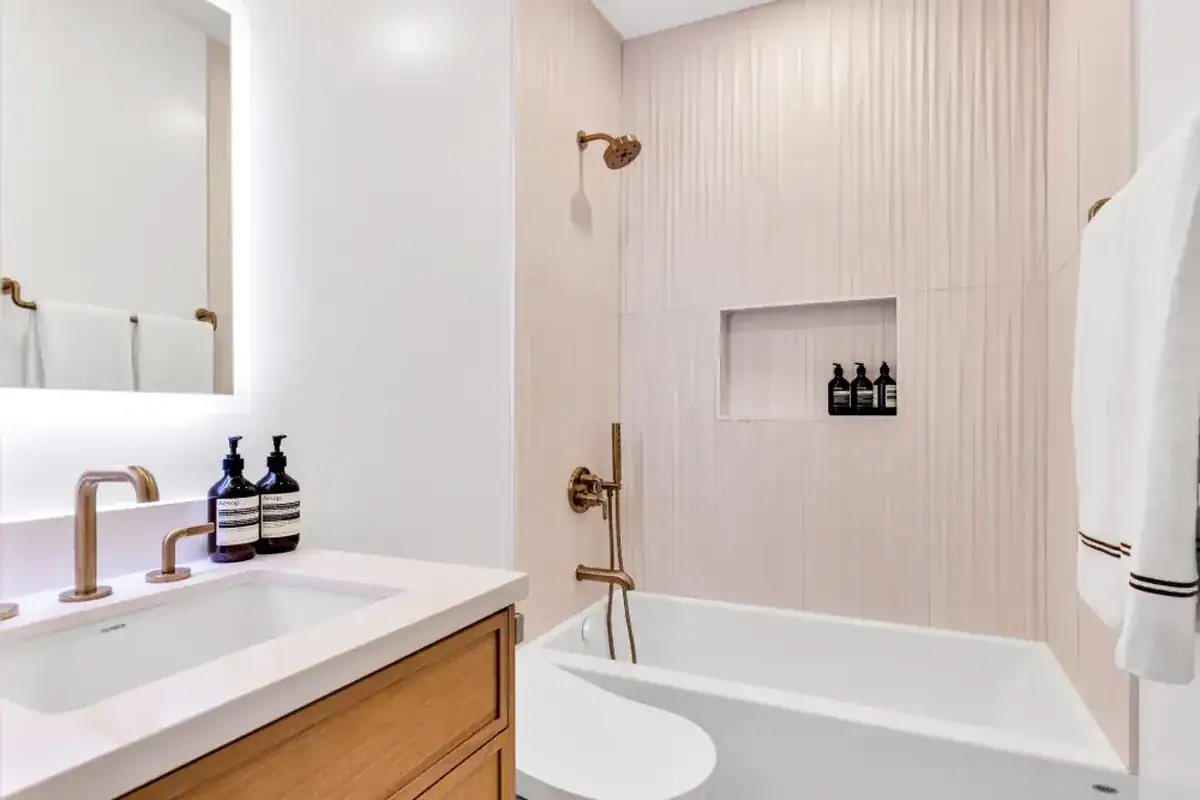 Small bathroom tub-shower with light textured vertical tile, a recessed shower niche, and a wood vanity with brass fixtures