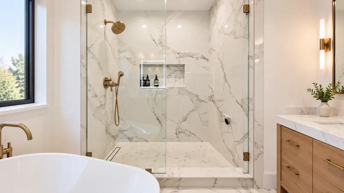 Walk-in shower with frameless glass doors, marble-look walls, linear drain, and brushed brass fixtures
